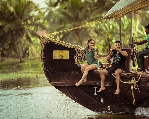 A happy couple wearing shorts and t-shirts and holding hands on a wooden boat in a lake surrounded by palm trees and hills during their Kerala tour package from Chennai. The image shows the beauty and romance of Kerala tourism.